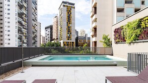Un área de piscina en la azotea con sillones de descanso, una piscina y edificios altos al fondo.