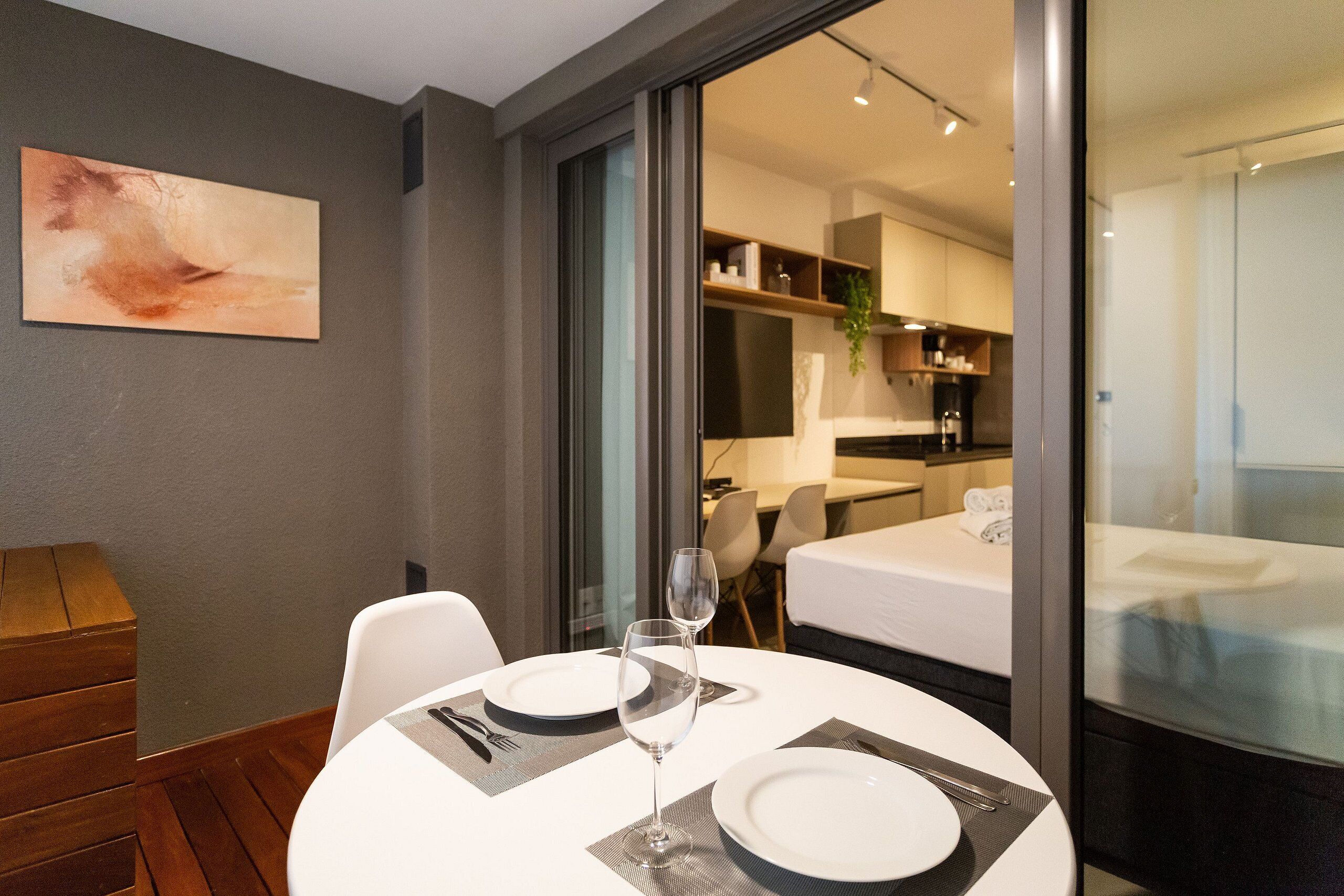 A dining area with a table set for two, a painting on the wall, and a view into the kitchen and bedroom through a glass door.