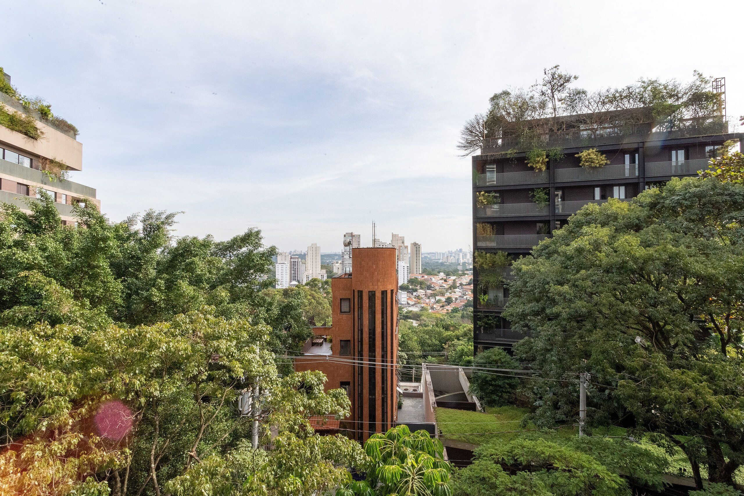 Un edificio moderno con una torre central, rodeado de abundante vegetación y otros edificios residenciales al fondo.