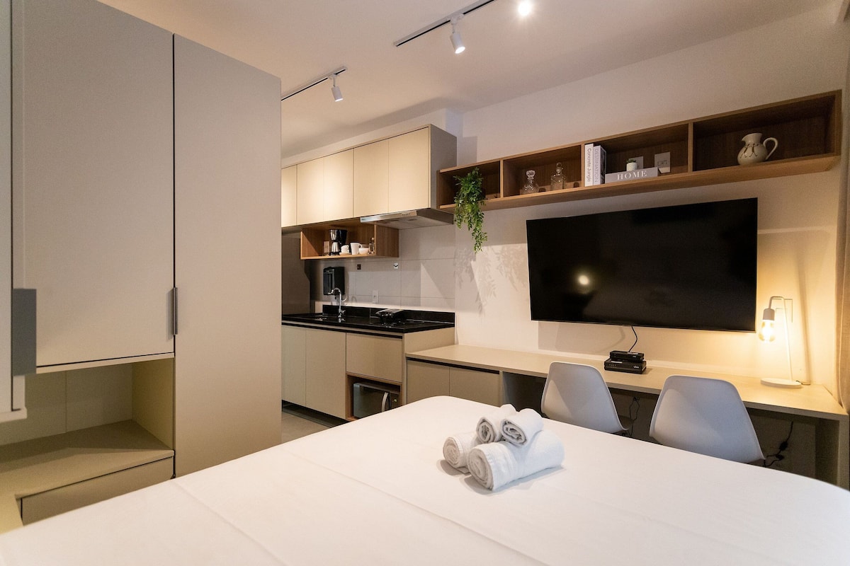 Una habitación de hotel moderna con una cama grande, un escritorio con sillas, un televisor de pantalla plana, una zona de cocina y un estante con objetos decorativos.