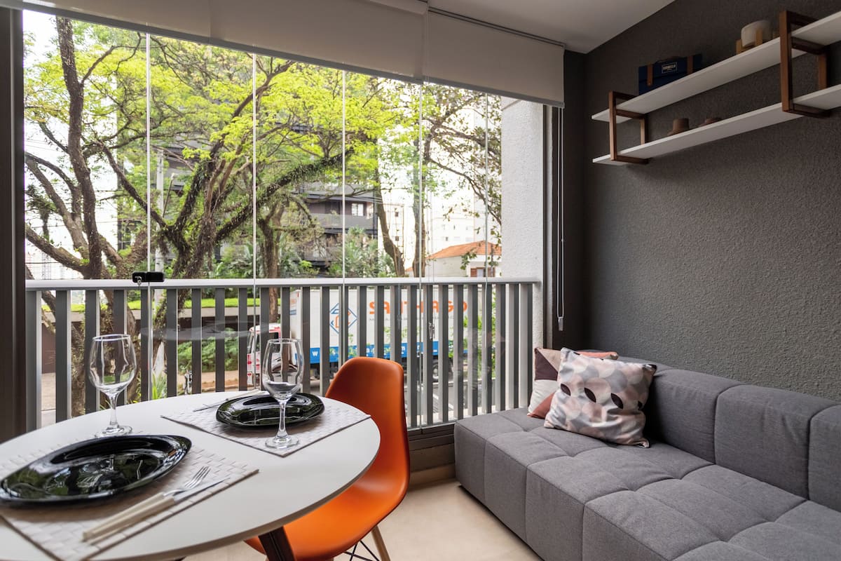 Una habitación moderna con balcón, un sofá gris, una mesa de comedor con sillas y vistas a árboles y edificios.