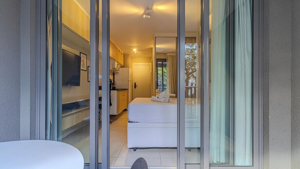 Una habitación de hotel moderna con puertas correderas de cristal que dan a un balcón, una cama, una kitchenette pequeña y un televisor de pantalla plana.