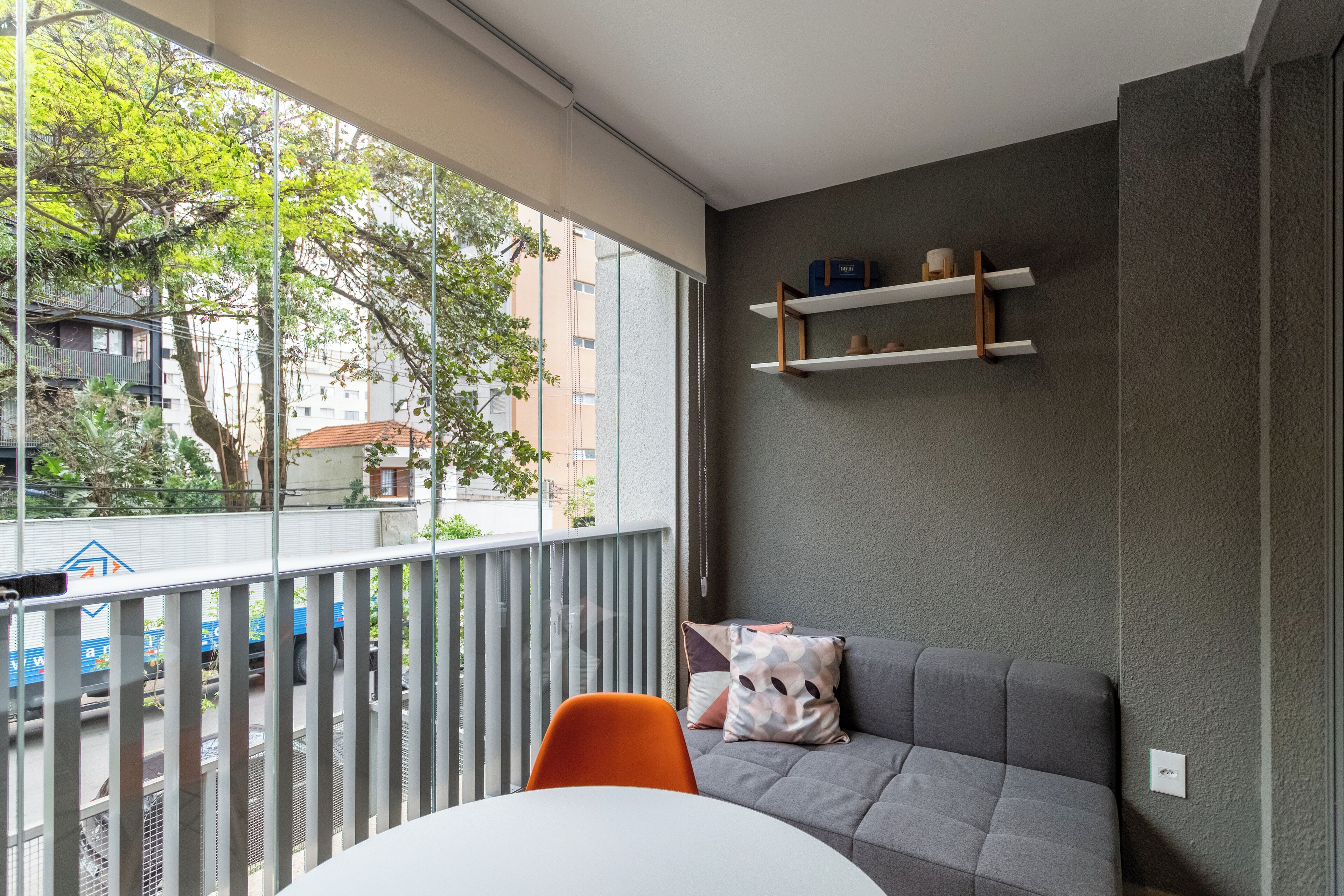 Un balcon donnant sur des immeubles et des arbres, un canapé gris avec des coussins, une table blanche et une étagère avec divers objets.