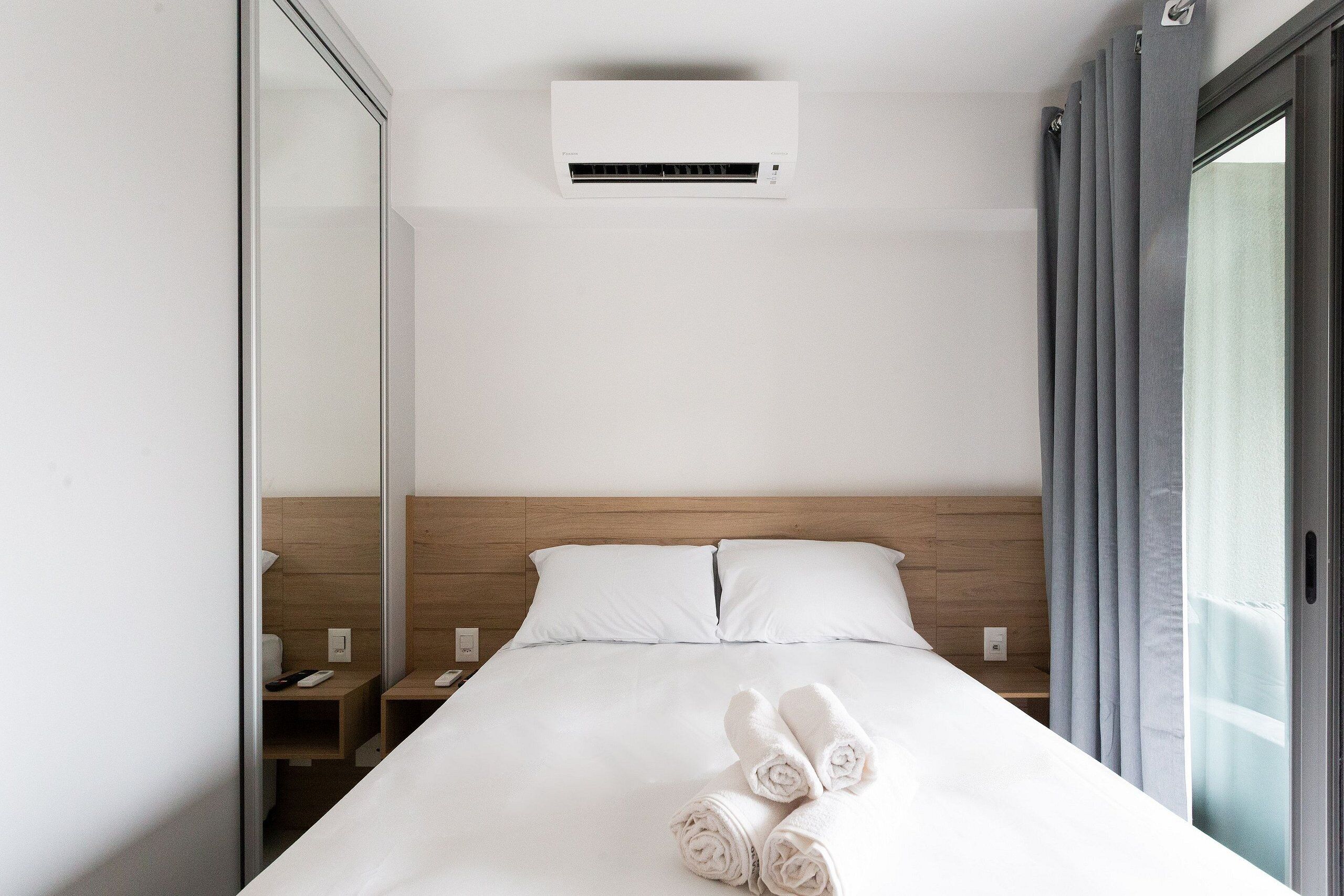 Et hotelværelse med en seng, et natbord, en vægmonteret aircondition og et vindue med gardiner.