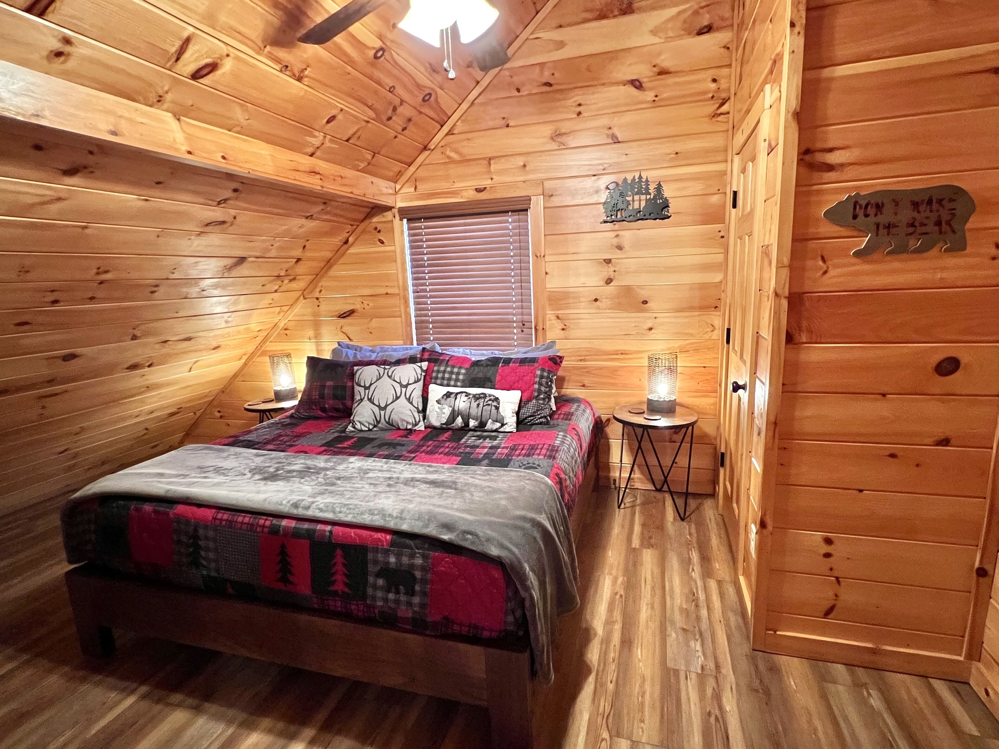 Un dormitorio de cabaña acogedor, con techo de madera, cama con cubrecama a cuadros, mesita de noche con lámpara y puerta con silueta de oso.
