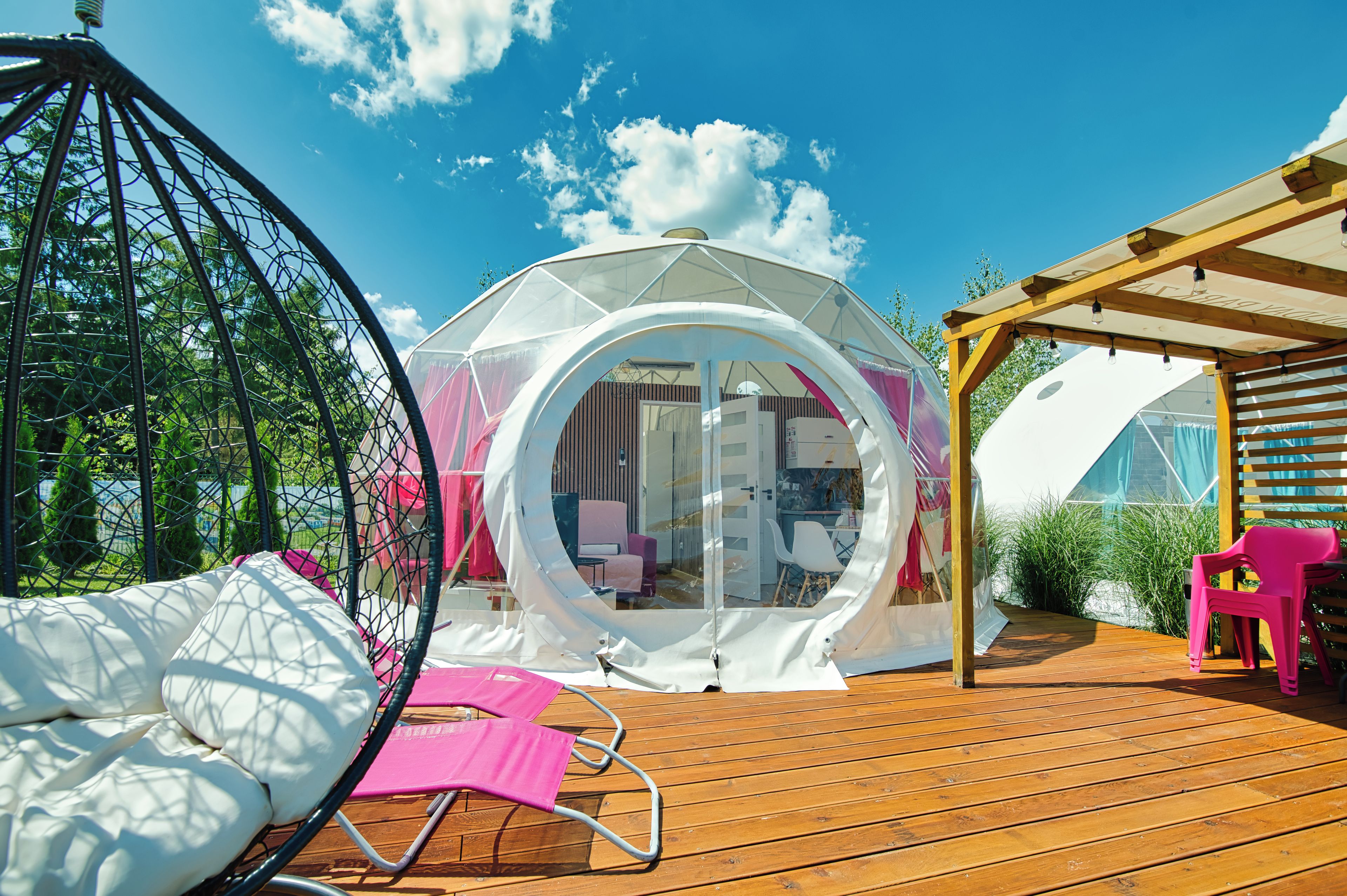 A transparent dome structure with a wooden deck, a hanging chair, and a pink chair.