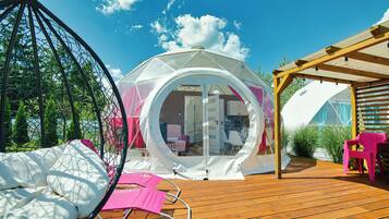 A transparent dome structure with a wooden deck, a hanging chair, and a pink chair.