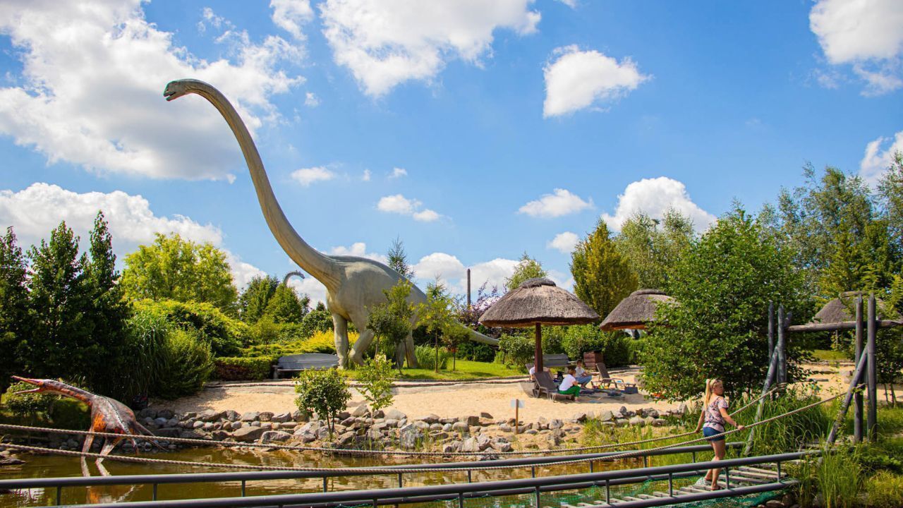 Exposição de dinossauros com um grande saurópode, um modelo de dinossauro pequeno e um lago.