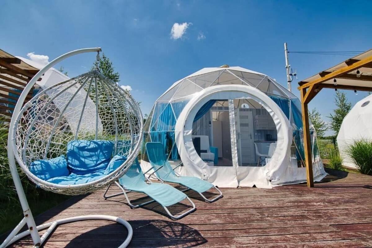 Une terrasse en bois avec un fauteuil suspendu, deux tentes blanches et une pergola.