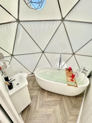 Een modern, geometrisch interieur met een ovaal bad in het midden, een klein wastafeltje en een houten dienblad met rode glazen.