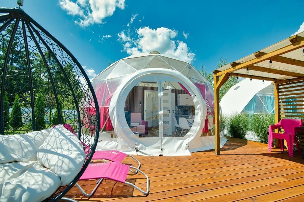 Uma estrutura em cúpula transparente com um deck de madeira, uma cadeira suspensa e uma cadeira rosa.