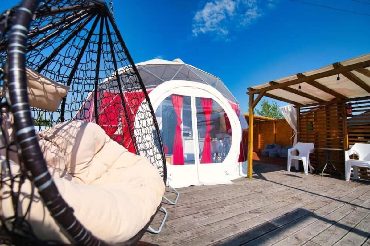 Un espace extérieur accueillant, avec une terrasse en bois, un fauteuil suspendu et un gazebo blanc agrémenté de rideaux rouges.
