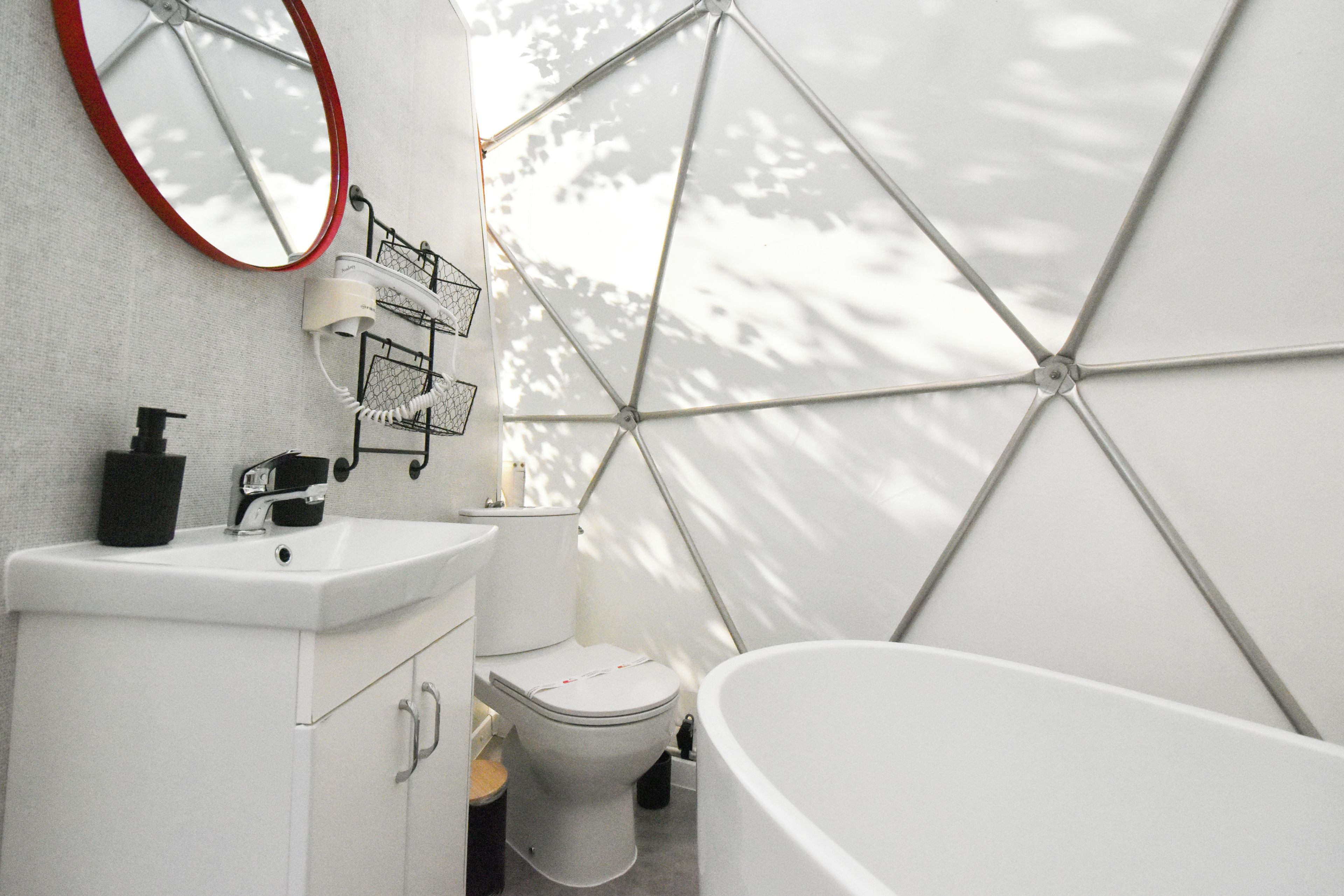 A modern bathroom with a white sink, toilet, and bathtub. There is a mirror with a red frame and a hairdryer on the wall.