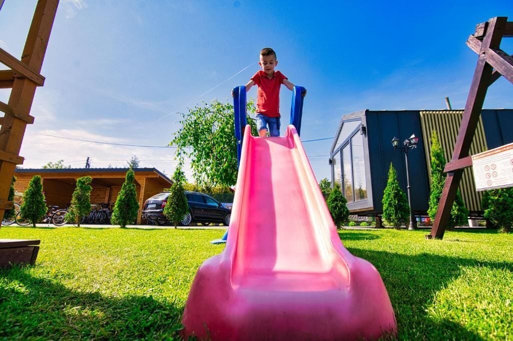 A child on a pink slide in a backyard with a wooden deck, a car, and a building in the background.