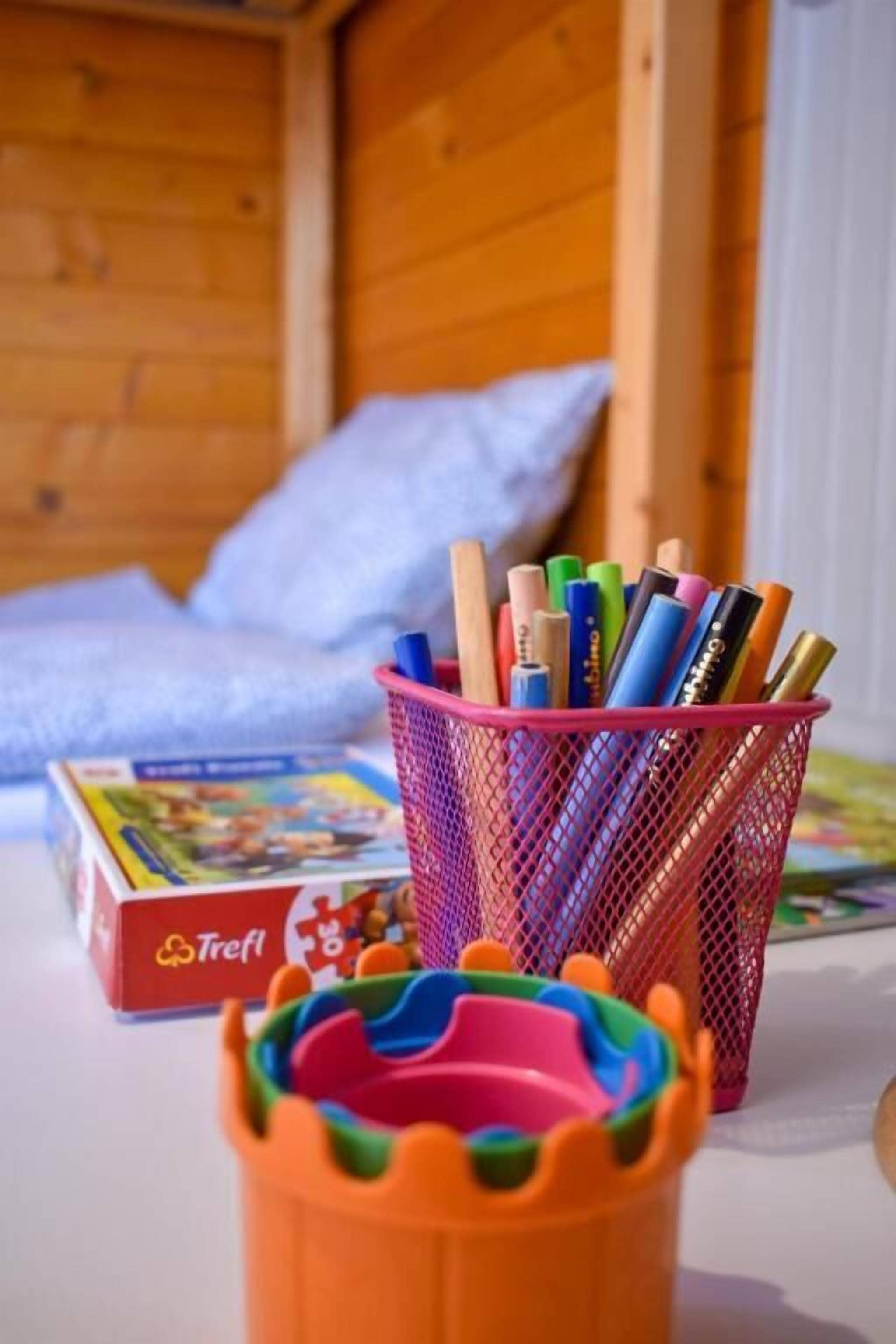 Ein Holzhütten-Interieur mit einem Bett, einem bunten Schulmaterial-Korb und einer Trefl-Puzzlebox.