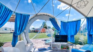 Un área de estar al aire libre con carpa, cortinas azules, un televisor y asientos.