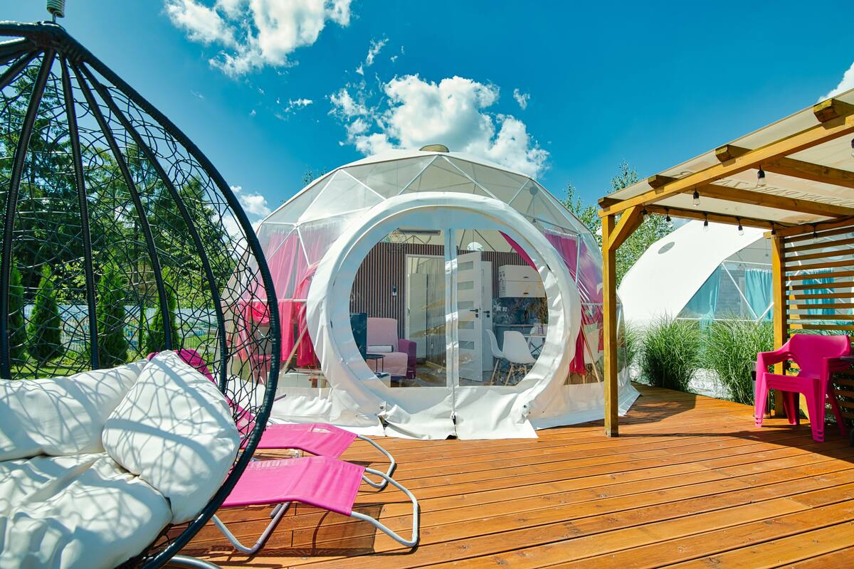 Une structure en forme de dôme transparent, dotée d’une terrasse en bois, d’une chaise suspendue et d’une chaise rose.