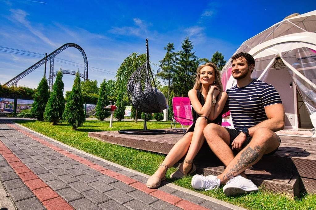 Ein Paar sitzt auf einer Bank in einem Park, im Hintergrund ist eine Achterbahn und ein Zelt zu sehen.