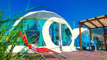 Uma estrutura em forma de cúpula com teto transparente, rodeada por um deck de madeira e uma área de estar coberta.