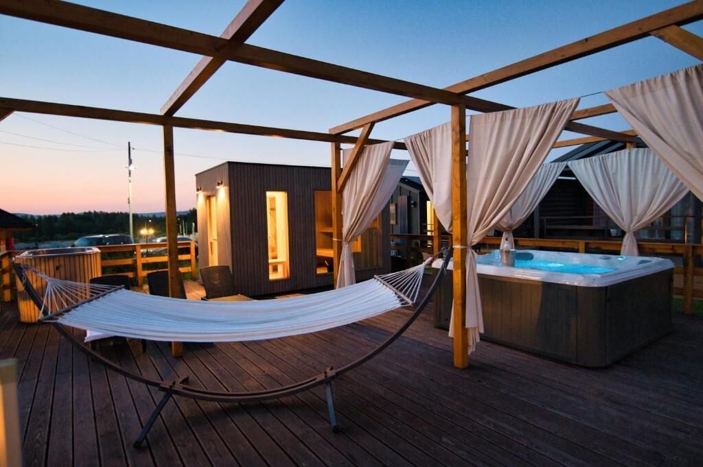Una terraza de madera con una hamaca, un jacuzzi y una pequeña construcción con ventanas.