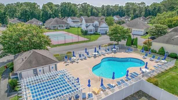 Un quartier résidentiel doté d’une piscine entourée de chaises longues et de parasols, d’un court de tennis et de maisons en arrière-plan.