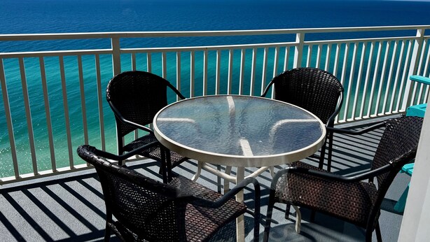A balcony with a round glass table and four wicker chairs, overlooking a turquoise sea.