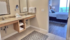 A hotel bathroom with a double sink, vanity, and a large mirror with lights. There is a towel rack and a wooden shelf with towels. The bathroom leads to a bedroom with a bed, a nightstand, and a view of the ocean.