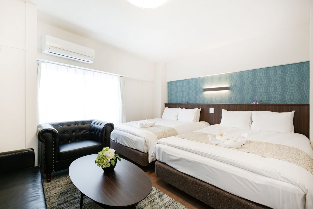 A hotel room with two beds, a black leather sofa, a round wooden table, and a large window.