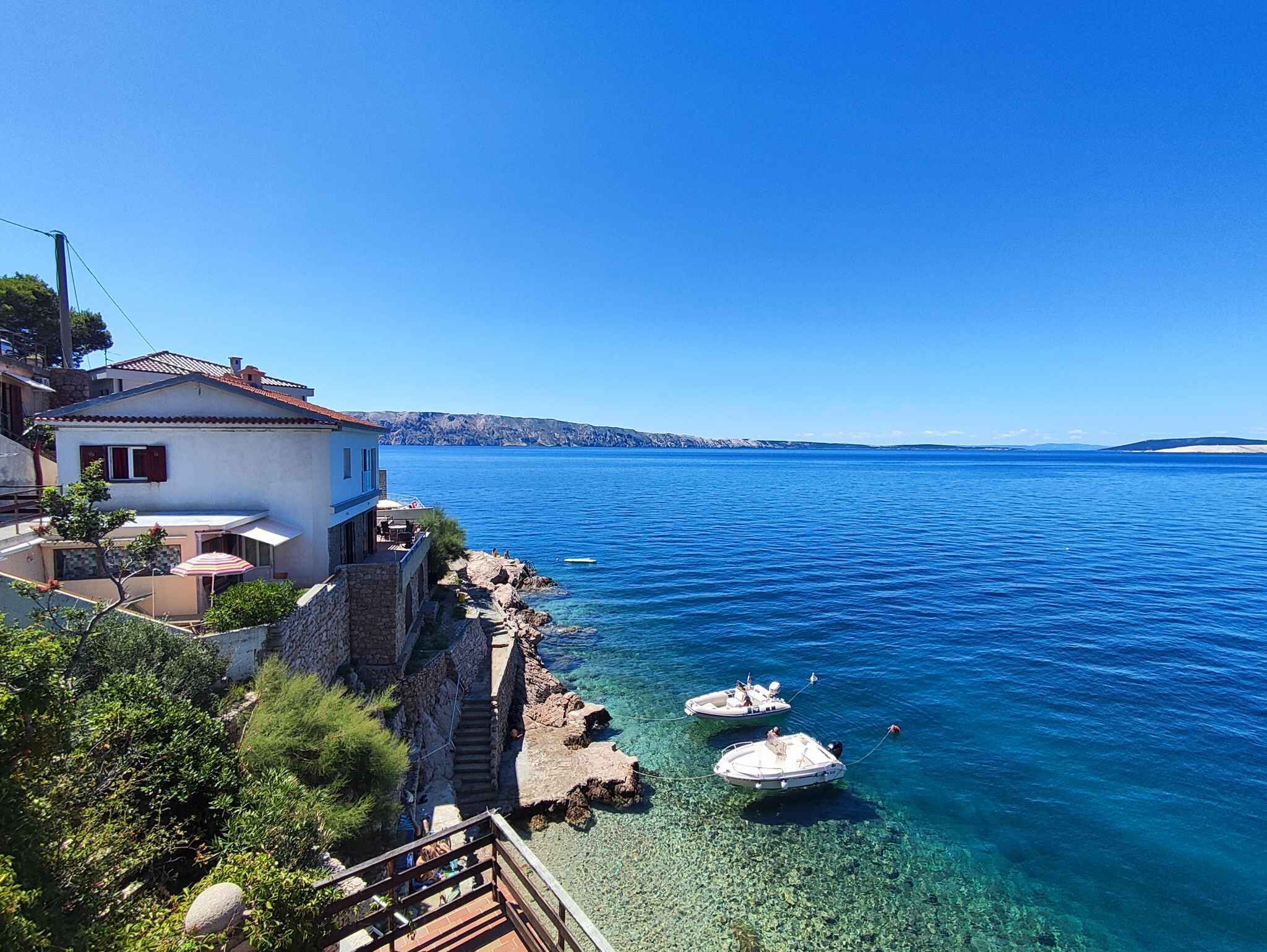 A coastal house with a balcony overlooking clear blue waters, boats anchored nearby, and a rocky shoreline.