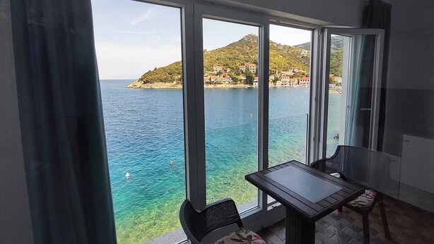 Un balcón con mesa y sillas, con vista a una zona costera de aguas azules cristalinas y un paisaje montañoso.