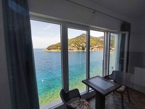 Un balcon avec une table et des chaises, donnant sur une zone côtière aux eaux d’un bleu limpide et un paysage vallonné.