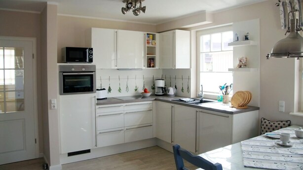 A modern kitchen with white cabinets, a microwave, oven, and a coffee maker. There is a dining table with a chair and a window with a view. - Semi-detached house - Isabella - Bright vacation home spread over 3 floors with (Zingst)