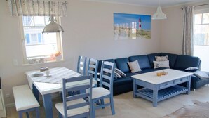 A living room with a blue sofa, white coffee table, and matching chairs. There is a dining table with chairs and a window with curtains. - Semi-detached house - Isabella - Bright vacation home spread over 3 floors with (Zingst)