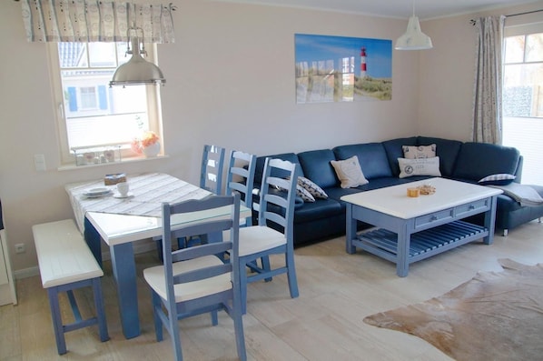 Ein Wohnzimmer mit einem blauen Sofa, einem weißen Couchtisch und passenden Stühlen. Es gibt einen Esstisch mit Stühlen sowie ein Fenster mit Vorhängen.