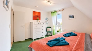 A bedroom with a bed, a dresser, a chair, a window with curtains, and artwork on the walls.