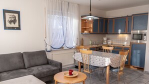Un espace de vie compact comprenant un canapé, une table à manger, des chaises, un coin cuisine avec des placards et des appareils électroménagers, et une fenêtre avec des rideaux.