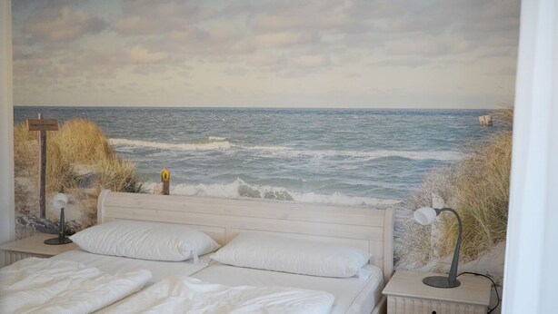 Une chambre avec une grande fresque murale représentant une scène de plage, un lit avec du linge de lit blanc, deux tables de chevet avec des lampes et une petite table de nuit avec une lampe.