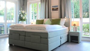A modern bedroom with a large bed, grey bedding, a bedside table with a lamp, and a view of greenery through large windows. - Modern 3-room apartment directly behind the Baltic Sea dike - Logierhaus Friedrich (Zingst)