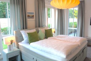 Ein ordentlich bezogenes Bett mit weißen Laken und grünen Kissen, ein Nachttisch mit Lampe sowie ein großes Fenster mit Vorhängen.