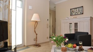 Ein Wohnzimmer mit Fernseher, Lampe, Sofa, Tisch mit Obst und Blumen sowie einer Vitrine mit Gläsern.
