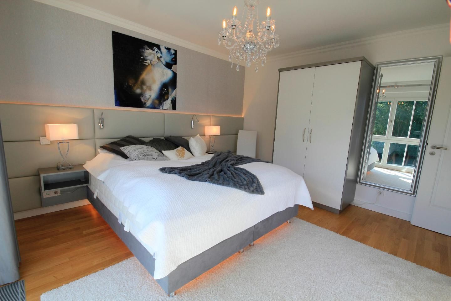 A modern bedroom with a large bed, bedside lamps, a chandelier, and a painting on the wall.