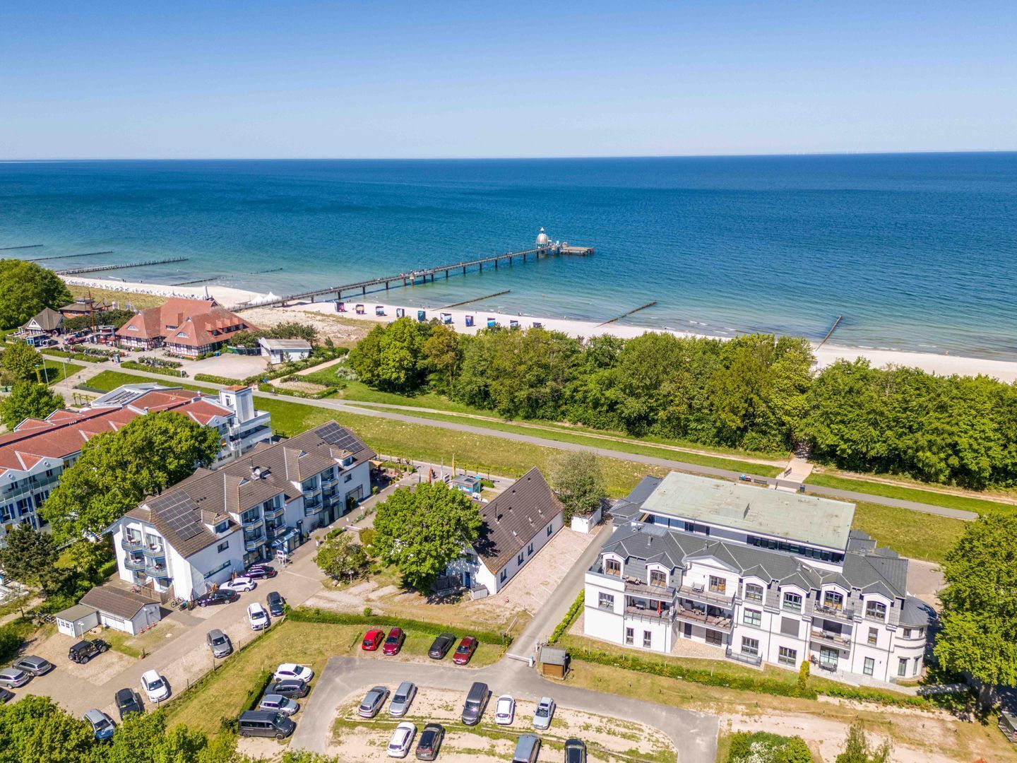 Vista aérea de una zona costera con un muelle que se adentra en el mar, una playa y un conjunto de edificios con aparcamientos.