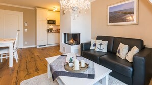 A living room with a fireplace, a black leather sofa, a dining table with chairs, and a chandelier. - Exclusive vacation apartment with limited sea view and balcony in Zingst - (Zingst)
