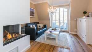 A living room with a fireplace, a black sofa, a coffee table, and a view of the outdoors through a large window. - Exclusive vacation apartment with limited sea view and balcony in Zingst - (Zingst)