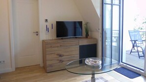 Una sala de estar moderna con un televisor sobre un mueble de madera, una mesa de vidrio con un cuenco y un ventanal corredero que da a un espacio exterior.
