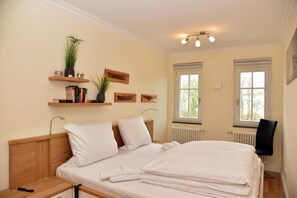 Une chambre avec une tête de lit en bois, un lit recouvert de draps blancs, une table de chevet, une commode, une chaise noire, une fenêtre donnant sur un paysage agréable et des étagères murales ornées de livres et d’une plante.
