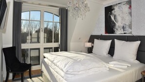 Une chambre d’hôtel avec un grand lit, un lustre, une chaise noire, une fenêtre avec des rideaux et un tableau au mur.