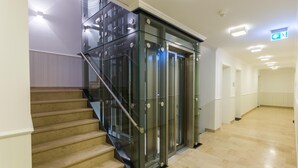 Un intérieur moderne doté d’un ascenseur en verre, d’un escalier et d’un couloir au revêtement de sol de couleur claire.