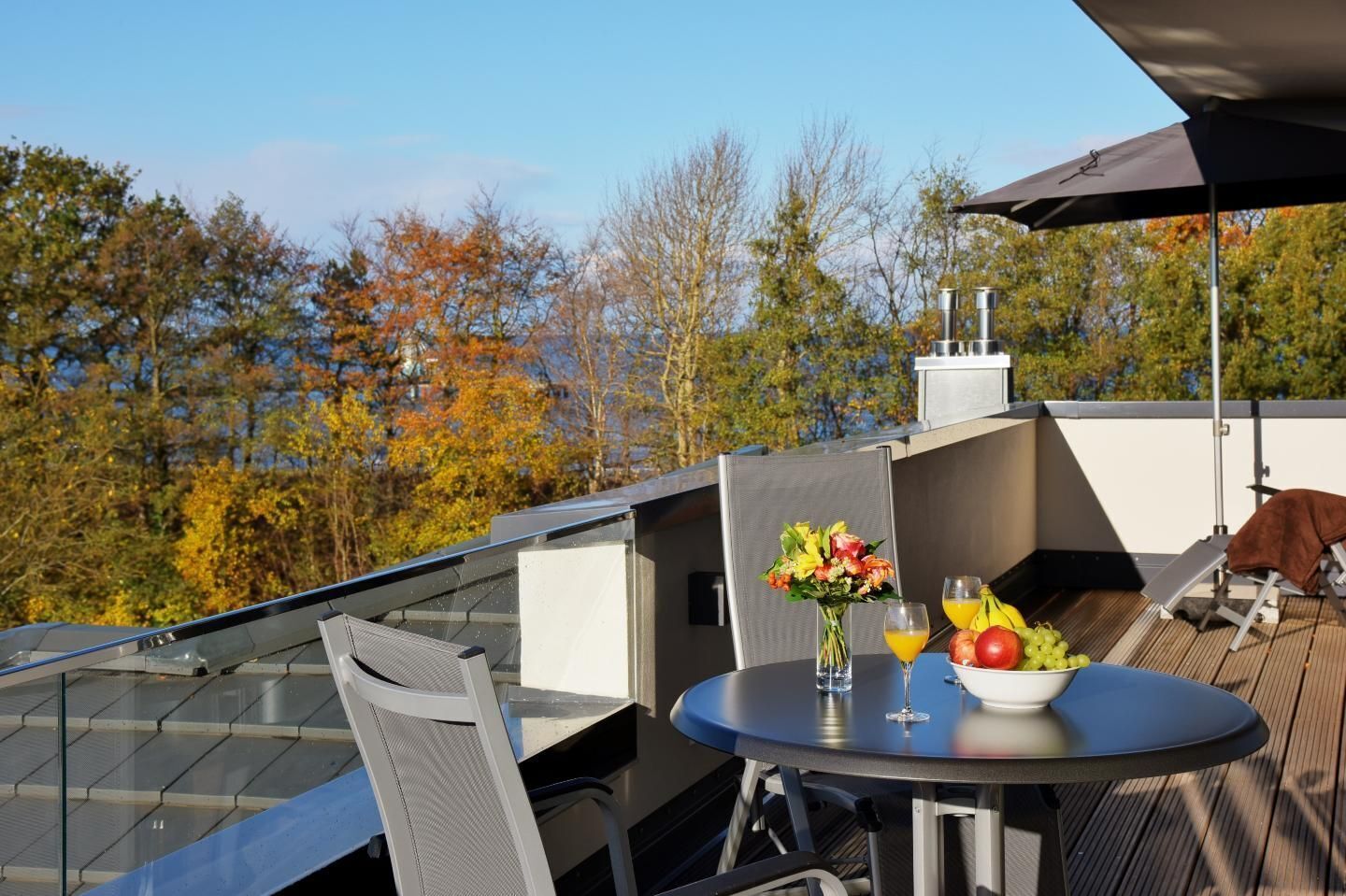 A rooftop terrace with a table set for two, a chair, a glass of orange juice, a bowl of fruit, and a vase of flowers.