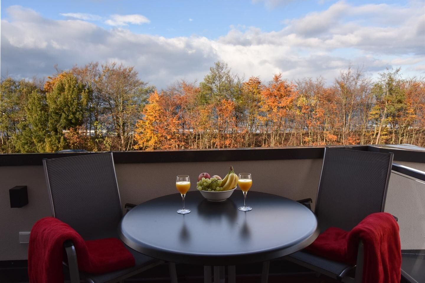 Une table ronde avec deux chaises, un saladier de fruits et deux verres de jus d’orange, le tout sur un balcon avec, en arrière-plan, le feuillage automnal.